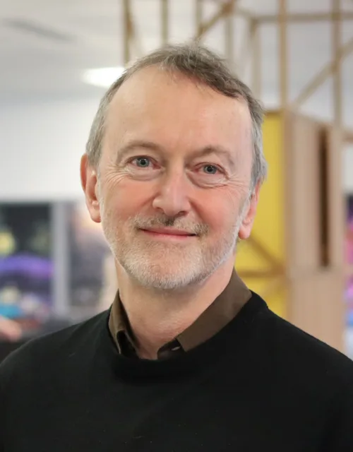 Alasdair smiling and looking at the camera in a brown shirt and black sweater in an office space