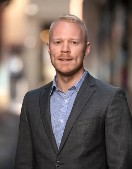 David stood in a blue shirt and grey suit jacket in a soft focus street setting