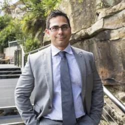 Mohammad Ali in a grey suit and blue shirt and tie with stairs and wall in the background